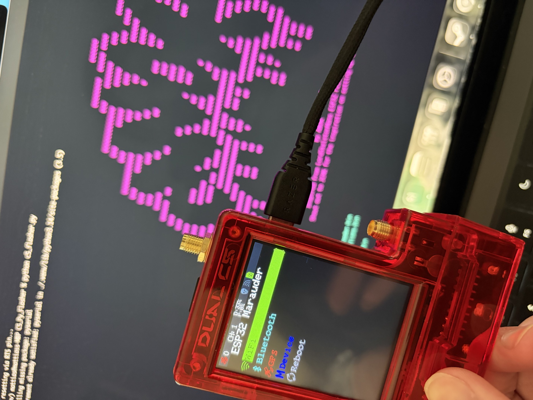 A custom Dual C5 ESP32 Marauder board in a translucent red case held in front of a monitor showing a flasher script and pink ASCII art output.