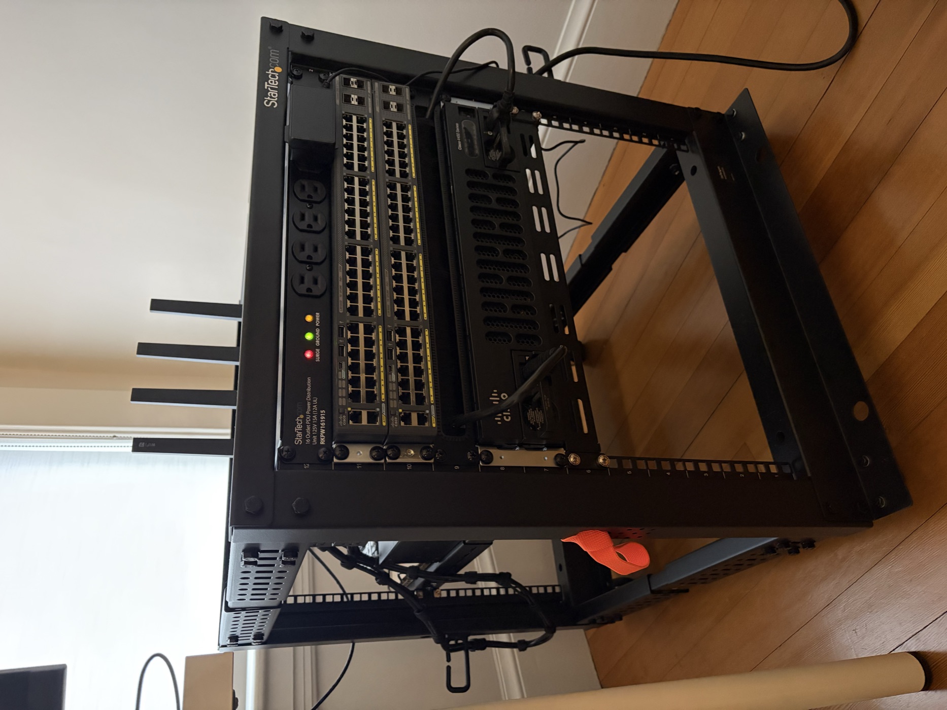 A black networking rack on a hardwood floor with a Cisco switch, patch panel, and StarTech PDU mounted on the front.
